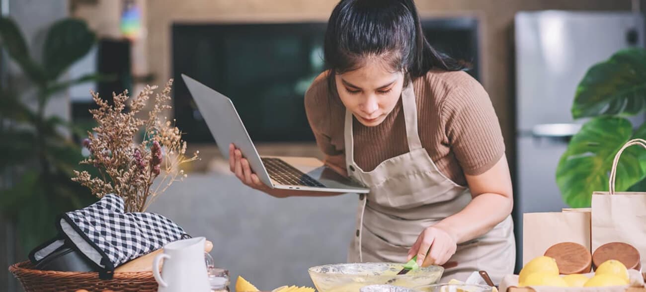 Woman preparing a meal at home while reading a plan on her laptop